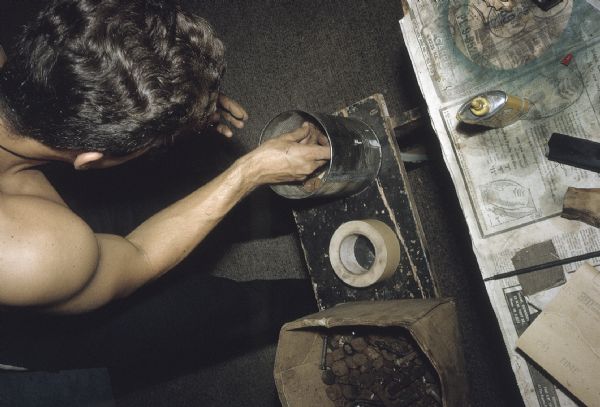 Overhead view of a member of the anti-Castro group Commandos L manufacturing explosive devices. The man is placing metal scrap such as rusty bolts and washers in a large can. A roll of masking tape, a sheet of sandpaper and a can of glue also sit on the bench and newspaper-covered worktable.