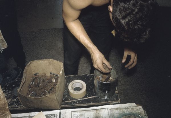 Front view of a member of the anti-Castro group Commandos L manufacturing explosive devices. The man is placing metal scrap such as rusty bolts and washers in a large can. A roll of masking tape sits next to the can on the bench.