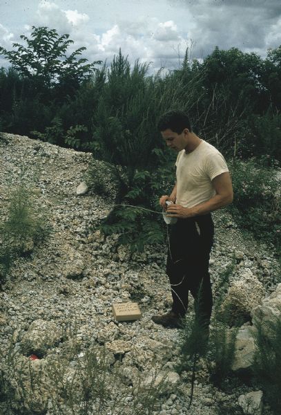 Member of the anti-Castro group Commandos L setting up to test an incendiary device in the Everglades. A man stands above a brown package in a gravel clearing holding a roll of wire.