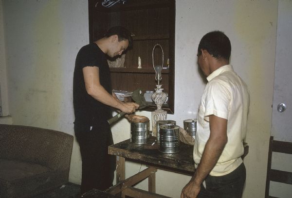 Two members of the anti-Castro group Commandos L making explosive devices at the group's barracks/armory. One member is hammering cans closed on a table while the other watches.