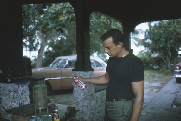 Member of the anti-Castro group Commandos L spray painting a homemade explosive device with a camouflage pattern under a covered porch. Parked cars and trees can be seen in the background.