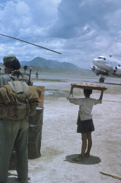 Airstrip at Luang Prabang airport, Laos. A young food vendor stands with a large circular tray on his head which casts shade for him. A silver plane sits on the dirt runway in the background and a few soldiers stand to the side.