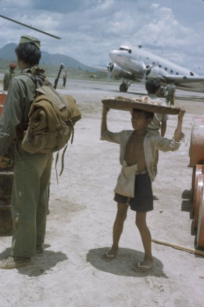 A young vendor stands with a large plate of food on his head and a satchel around his neck at the runway of the Luang Prabang Airport, Laos. There is a silver plane in the background and a few soldiers are  standing on the left.