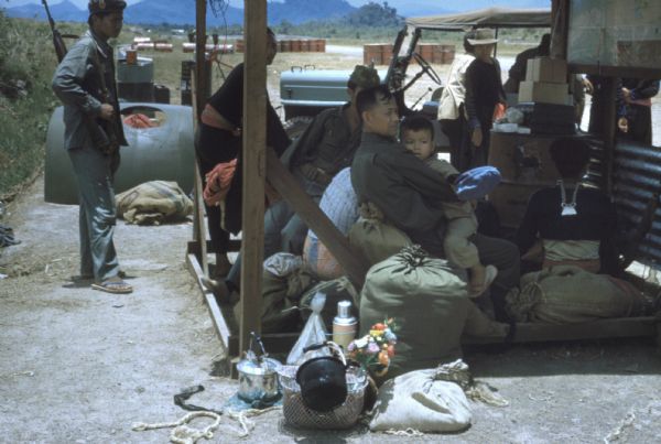 A small group of refugees wait with luggage under an awning at Luang Prabang airport, Laos. An armed soldier stands behind them on the right. There is a truck in the background.