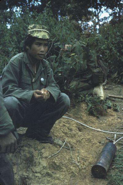 Colonel Bounchon's Lao infantry troops laying explosives forward of lines in the vicinity of Luang Prabang. Two soldiers crouch near some bushes with a metal canister near them in a low trench on the ground.