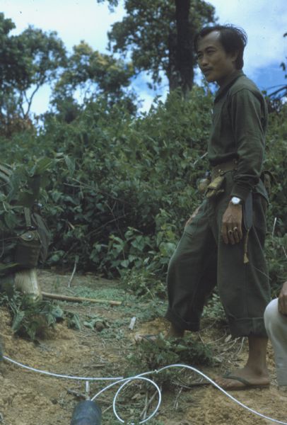 Colonel Bounchon's Lao Infantry laying explosives forward of lines in the vicinity of Luang Prabang, Laos. A soldier stands in a clearing in a wooded area. There is a metal canister with a line running from it at his feet.