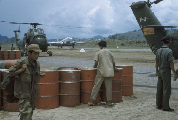 Gas drums for fueling helicopters at the Luang Prabang airport, Laos.  A few soldiers are in the foreground, and two helicopters and an airplane are on the runway.