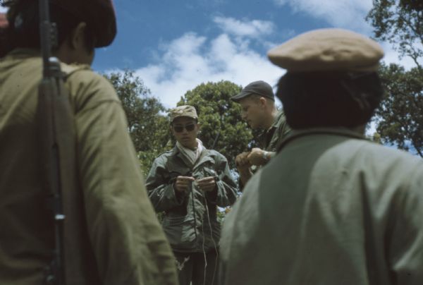 Colonel Bounchon's Lao Infantry in the field in the Luang Prabang vicinity during a mission to lay explosives behind lines. Two men stand in the background holding wires, while two men in the extreme foreground watch them with their backs to the camera.