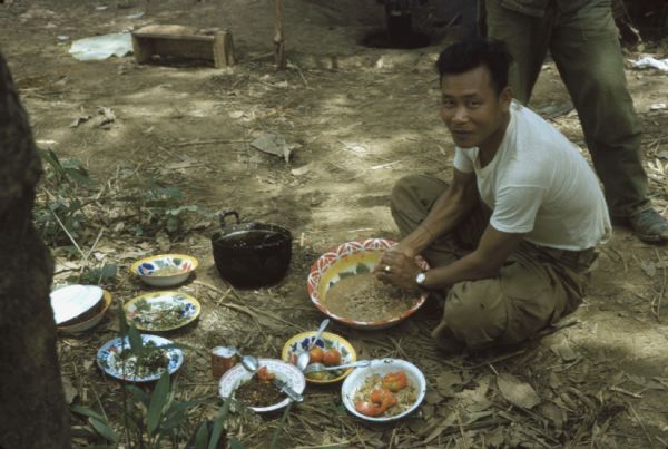 Colonel Bounchon's Lao Infantryman preparing a meal in a clearing in the Luang Prabang vicinity, Laos. A number of brightly-colored plates and bowls with food in them are sitting on the ground.
