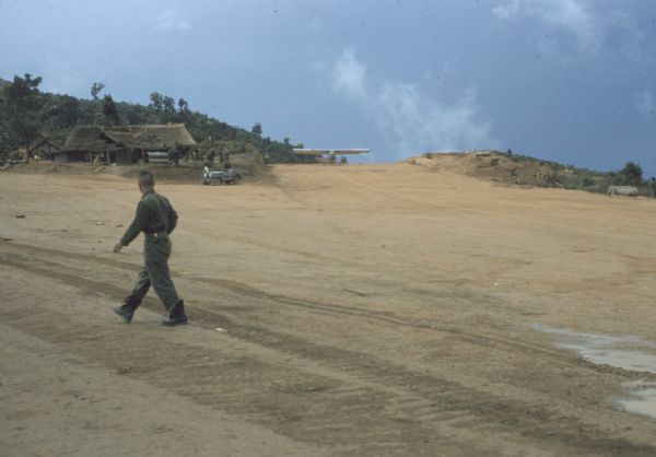 The wide dirt expanse of the Luang Prabang airport runway, Laos. A man is walking across a runway, and a plane is on another runway near a wooden building on a rise in the background.