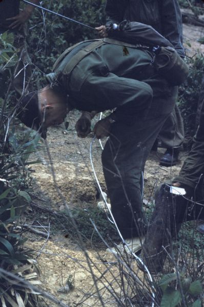 Colonel Bounchon's Lao Infantry unit laying explosives forward of lines in the vicinity of Luang Prabang, Laos. A man is bent over working on a silver wire. A few men stand next to him observing and holding materials.