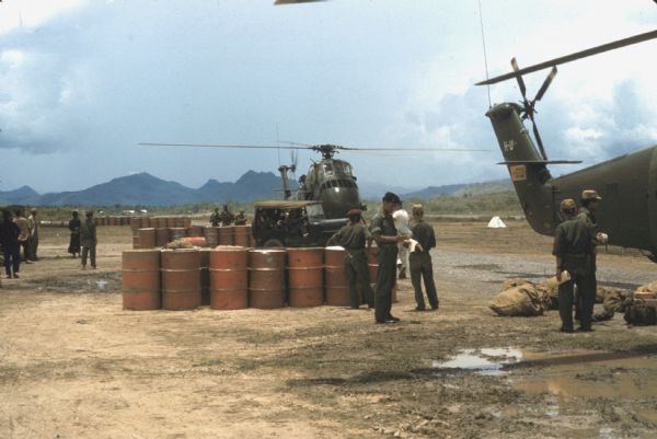 Helicopter refueling area at the Luang Prabang airport, Laos. Rows of orange 55-gallon drums are grouped at intervals on a dirt landing surface. Two helicopters sit on the ground and a number of soldiers are standing around. A full jeep is parked near a helicopter and mountains are visible in the distance.