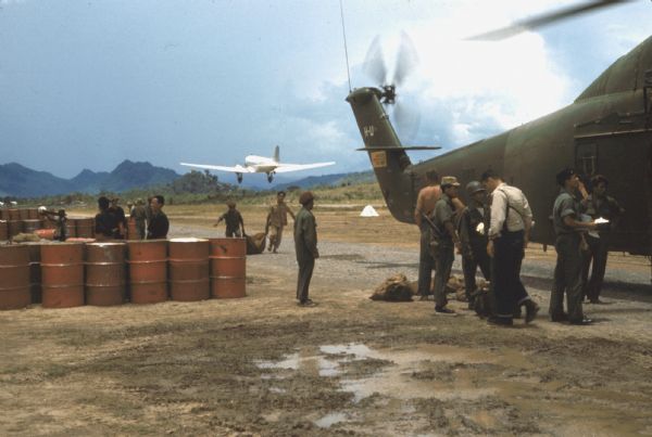 Helicopter and soldiers at the refueling area, Luang Prabang Airport, Laos. Soldiers stand next to the helicopter on the dirt landing surface. Others are working or standing in the background near 55-gallon drums of helicopter fuel. A silver plane has just taken off in the background.