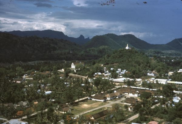 Aerial view of the town of Luang Prabang, Laos. Trees are interspersed among houses and roads and green mountains encircle the town. The golden roof of Phousi temple is visible on one hilltop.