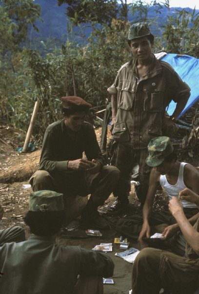 Group of Colonel Bounchon's Lao infantrymen playing cards in the field. The soldiers sit or stand around a blanket where cards, money and cigarettes are laid out. Green mountains are visible in the background.