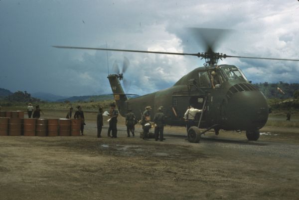 Loading of a military helicopter at Luang Prabang airport. A number of soldiers stand next to the helicopter which has its door open. 55-gallon drums of fuel are standing nearby on the dirt landing surface.