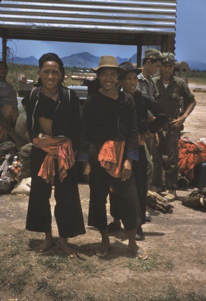 Three refugees posing for a picture in front of a corrugated structure for shade at the Luang Prabang airport, Laos. Two soldiers stand in the background and piles of personal belongings are clustered around.