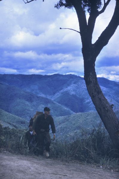 Colonel Bounchon's Lao Infantry men on mission to lay explosives behind lines. Three men are hiking up a hill. Rolling green mountains are visible in the background, and a tree stands by the trail.