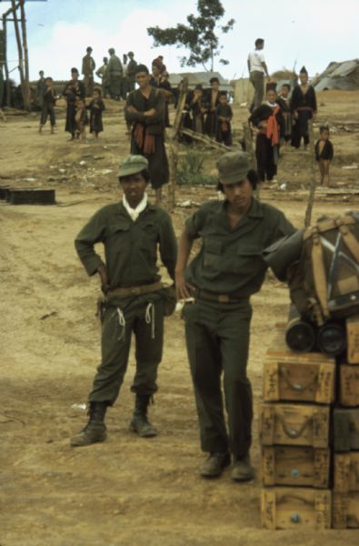 Two Lao infantry men posing for a picture on a dirt road next to a stack of crates in the Keukacham Meo settlement, Laos. A number of residents and soldiers are standing in the background across the road.