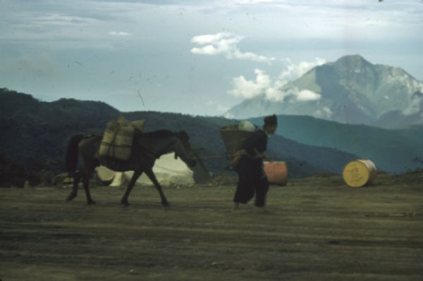 A woman carrying a basket on her back leads a mule laden with bundles across an airstrip at Keukacham village, Laos. There are 55-gallon drums near a tent, and mountains rise in the background.