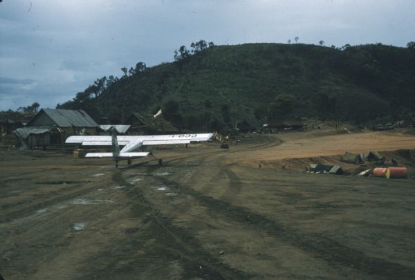Small plane sitting on Keukacham airstrip, Laos. A green hill rises in the background and tents and ramshackle buildings are on either side of the strip.
