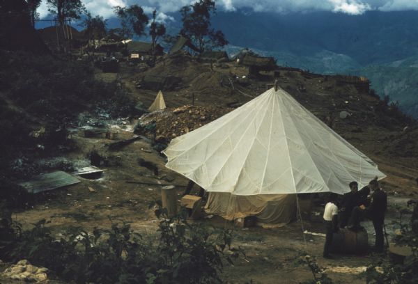 Elevated view of Keukacham village, Laos. Three soldiers stand talking in the foreground by a large, open, white tent. In the background is a rubble pile, more tents and thatched-roof buildings. The village overlooks rolling valleys and mountains.