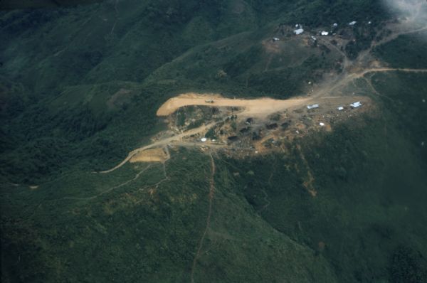 Aerial view of Keukacham village, Laos including landing strip with helicopter. The small settlement is clearly visible contrasted against the verdant green of the surrounding mountainside.