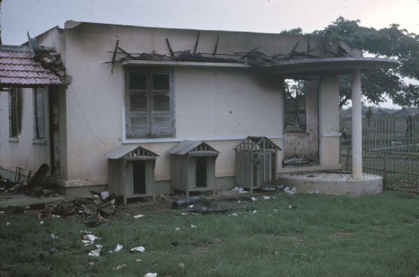 Building damaged by Viet Cong in Vinh Quoi, Vietnam. The pink building seems to have lost its roof and there is debris strewn about the yard. There are men in the far background.