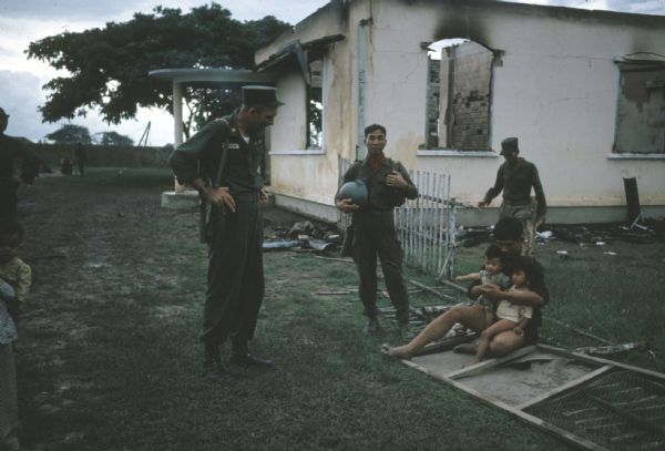 Vinh Quoi, Vietnam, in the aftermath of a Viet Cong attack. There is a burned-out building in the background. A male resident is sitting on a downed fence holding two children in his lap. A soldier is talking to him, and two other soldiers are standing nearby.