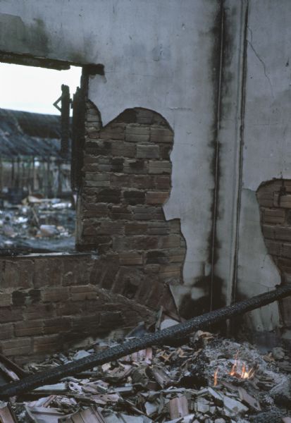 Interior of a wrecked building in Vinh Quoi, Vietnam, after a Viet Cong attack. Plaster from the walls and pieces of the roof form a heap of charred rubble on the floor which is still partially burning. Wreckage outside can also be seen from the opening where a window used to be.