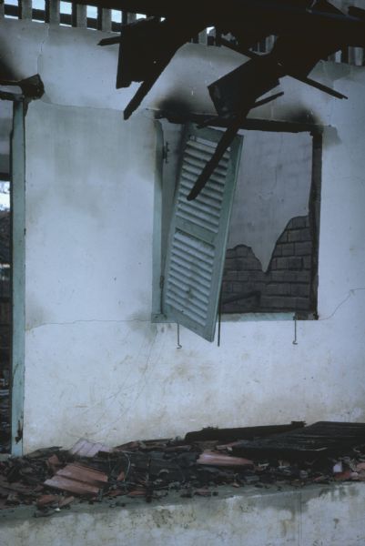 Interior of a window on a wrecked house in Vinh Quoi, Vietnam after a Viet Cong attack. One shutter hangs askew from the otherwise burned out window. Charred fragments of the roof and other shutters lie below on a ledge.