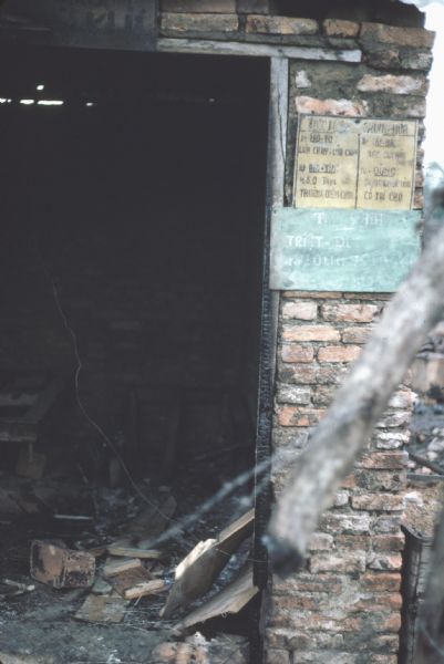 Entrance to a Vinh Quoi, Vietnam, building burned out in a Viet Cong attack. Debris can be seen cluttering the floor in the darkened interior. A tree branch scorched in the fire is in the foreground of the shot. Colored signs hang on the brick facade.