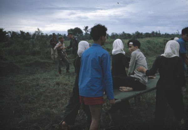 Vietnamese man and survivor of a Viet Cong attack on the village of Vinh Quoi is carried on a stretcher to a helicopter for medical evacuation. Three women wearing headscarves accompany the stretcher. Soldiers are talking in the grassy background.