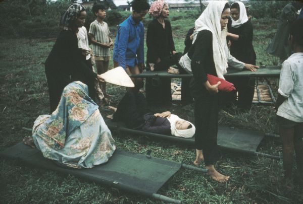 Survivors of a Viet Cong attack on the village of Vinh Quoi wait on or with stretchers for a helicopter medical evacuation in a grassy field.