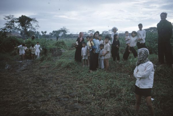 Women, children and elderly survivors of a Viet Cong attack on the village of Vinh Quoi standing in a field. In the background a group of men are hauling something on a blanket and some village buildings can be seen in the distance.