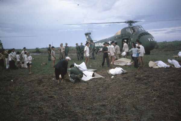 Helilift of corpses after a Viet Cong attack on the village of Vinh Quoi. The corpses are wrapped in sheets and lying atop mats on the ground in a field. Soldiers stand around a Marine helicopter and villagers stand in groups with the dead or nearby.