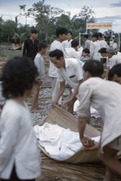 Aftermath of a Viet Cong attack on the village of Vinh Quoi. Children watch as a group of men begin to pick up corpses wrapped in sheets on woven mats.