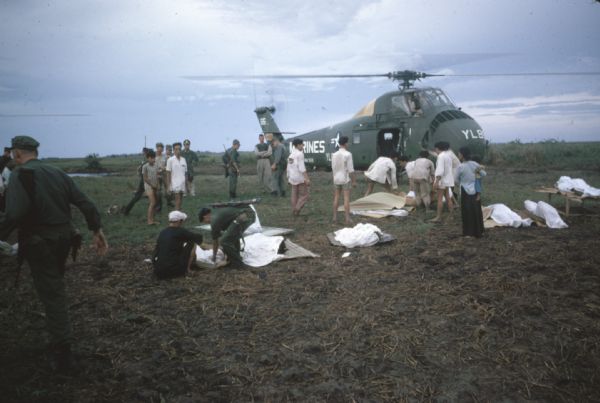 In the aftermath of Viet Cong attack on the village of Vinh Quoi, corpses await helilift. The corpses are wrapped in sheets and lying atop woven mats in a field next to a helicopter. Some of the corpses are being picked up while other soldiers and villagers watch.