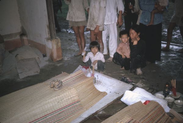 Funeral services in the village of Vinh Quoi, Vietnam for casualties of a Viet Cong attack. Two of the deceased are covered with woven mats and lying on white pallets. Bundles of incense are burning at the ends of the pallets and bunches of bananas sit on top of the mats. A woman and two children squat in front of one of the pallets and a small group of other people are standing behind them.