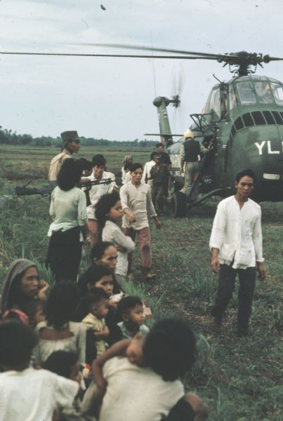 Survivors of a Viet Cong attack on the village of Vinh Quoi, Vietnam sit or stand near an American military helicopter that has arrived to offer aid. A soldier with a large gun is standing among them and other soldiers are near or in the helicopter.