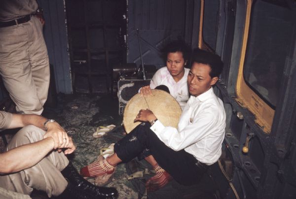 Two civilian survivors of a Viet Cong attack on Vinh Quoi, Vietnam sitting inside an American military helicopter that arrived to offer aid to the village. The civilians are sitting on the floor leaning up against a bare metal wall. The legs of two Americans also riding on the flight are visible from where they sit and stand on the other side of the craft.