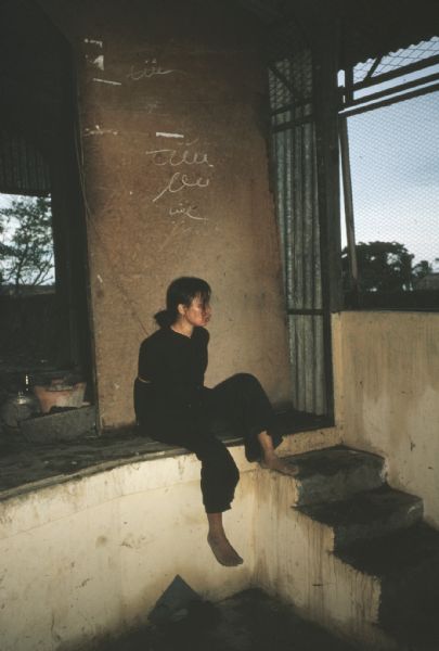 Captive held in a concrete room in Vinh Quoi, Vietnam in the aftermath of a Viet Cong attack on the village. The woman is wearing all black, is barefoot and has her arms bound behind her back. She is sitting on a concrete ledge in the bare room.