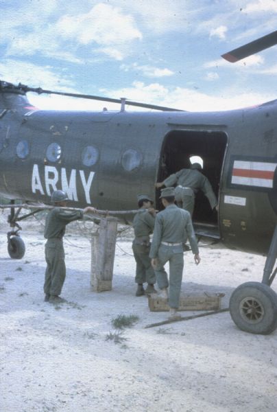 Troops load ammunition and rice delivered by plane to a forward airstrip into an Army helicopter in the Da Nang vicinity of Vietnam. Two of the men are carrying a large branch over their shoulder to carry an ammunition box.