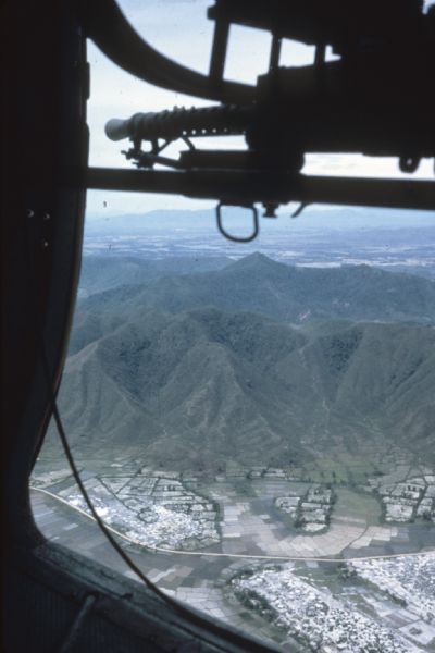 Silhouette of a machine gun in an Army helicopter and a scenic view of the mountain region in Vietnam below. Green mountains rise next to developed countryside with a patchwork of agricultural fields.