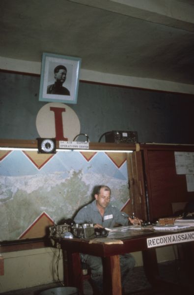 Soldier at work at a desk in an Air Support Operations Center, Da Nang, Vietnam. There is a large map on the wall, military radios, a clock and a military portrait in the Center.
