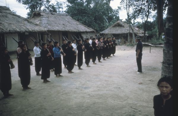 Women train with rifles in a village near the Laotion border in the mountain region of Vietnam. A group of about 25 women are lined up in two rows standing with rifles over their shoulders. A man stands in front of them. Thatch-roofed buildings are in the background. A girl is standing by a tree in the foreground.