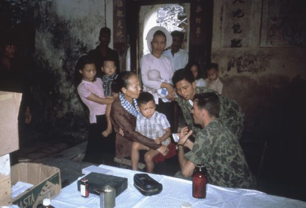 Joint MEDICO/U.S. Army humanitarian medical treatment mission to a forward village near the Laoation border in the mountain region of Vietnam. A man in fatigues is crouching on the floor of a stone building talking to a woman holding a boy while another man in fatigues listens. There is a table with medical supplies on it in the foreground and other women, children and men look on in the background. Mission was made by helicopter under fire most of the way.