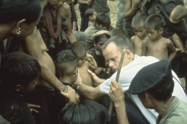 Medical examination of a boy in a refugee camp in the mountain region of Vietnam on the border of Laos. Treatment provided by a joint MEDICO/U.S. Army humanitarian medical mission. A man is examining the torso of a boy while a woman holds the child's shoulders. A group of other women and children crowd around.