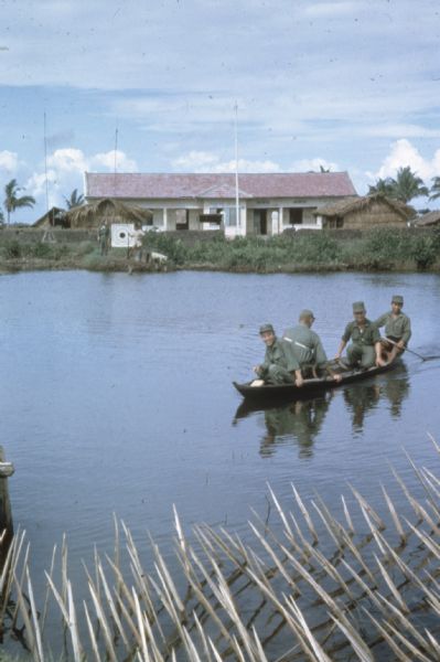 U.S. Army advisor Major M.C. Curley and soldiers crossing a river in the delta region of Vietnam in a small boat to visit an outpost. A large building, a marksmanship target and a few thatch structures are visible in the background on the opposite shore of the river.