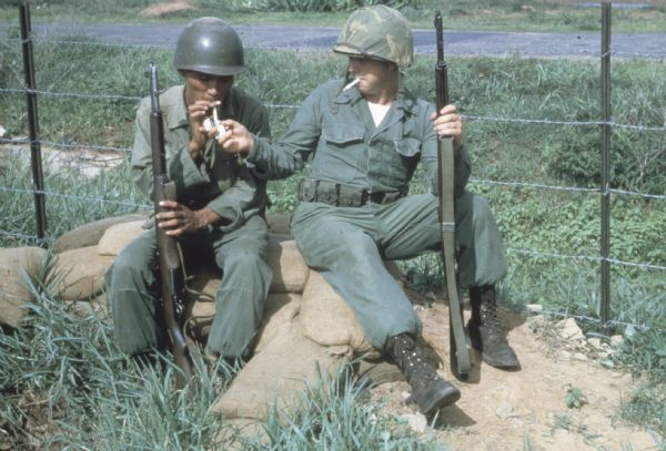 Two soldiers under the command of U.S. Army advisor Major M.C. Curley in the delta region of Vietnam sitting on sandbags smoking cigarettes.  The soldiers are holding their rifles, wearing helmets and sitting next to a barbed wire fence.